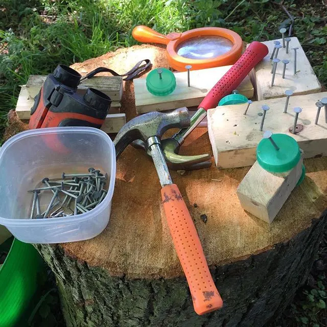 Garden tools and equipment arranged on a tree stump, including a hammer, magnifying glass, and containers.