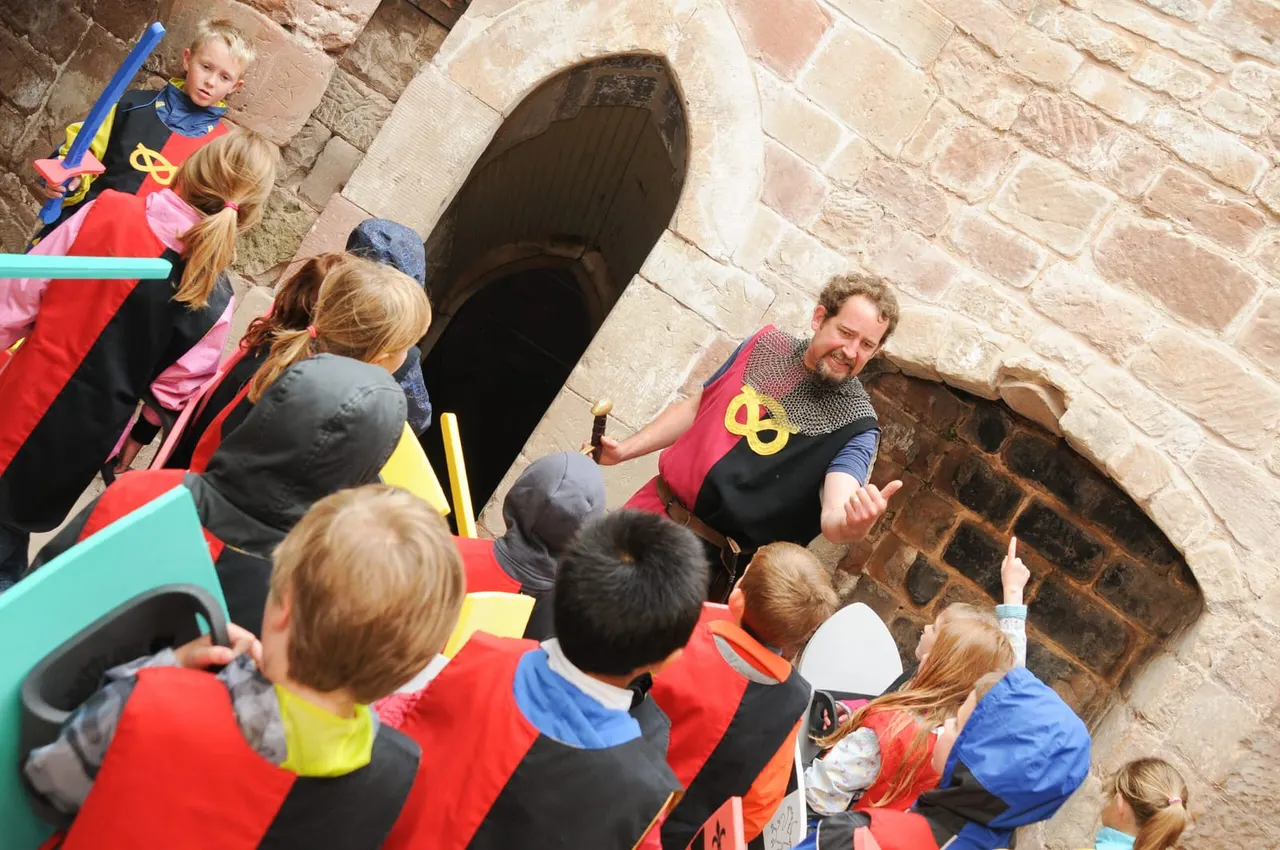 Adult guide in medieval costume addressing a group of children in fancy dress outside a stone castle archway.