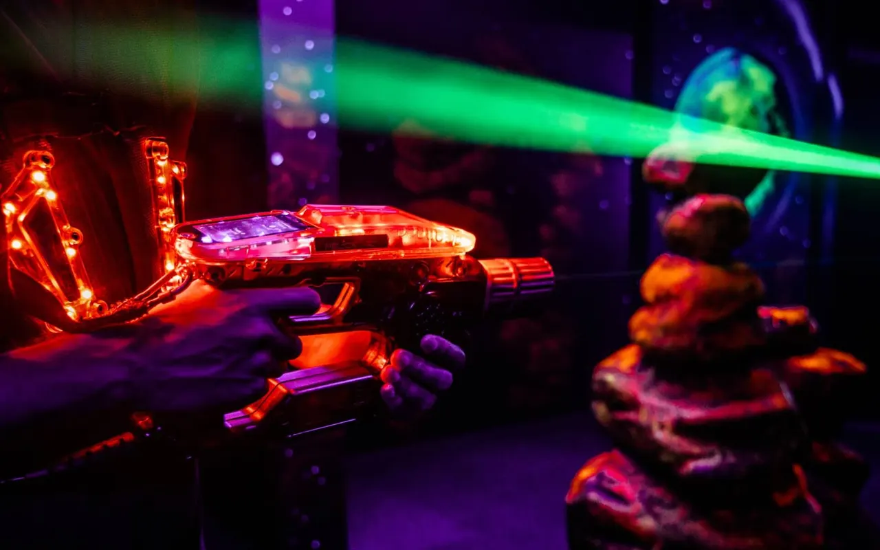 Players in a neon-lit laser tag arena holding illuminated laser guns with green laser beams.