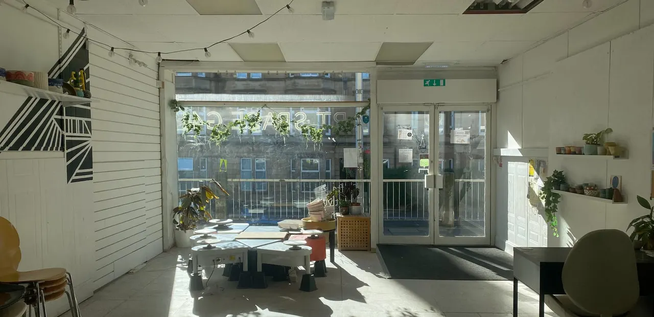 Modern art gallery interior with low tables, potted plants, and large windows overlooking a street.