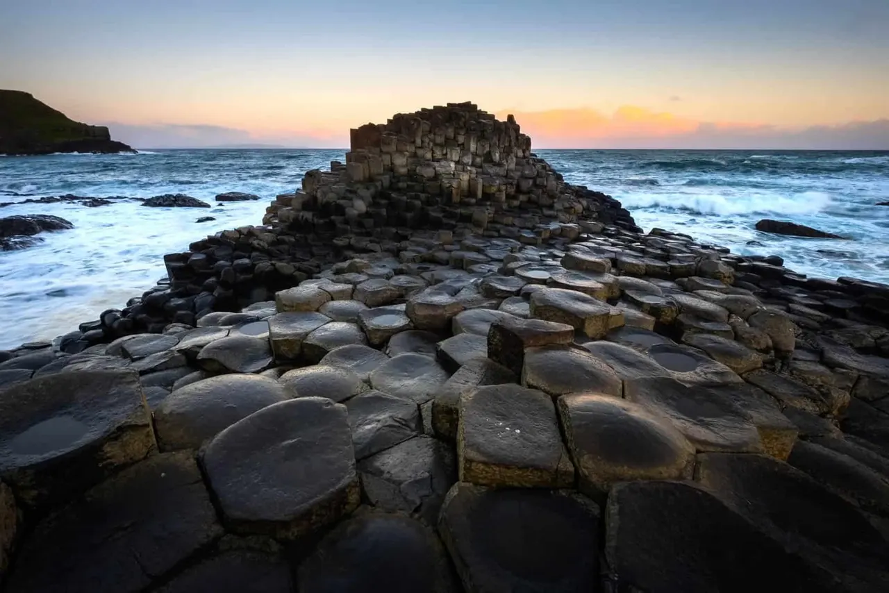 Hexagonal basalt columns on a rocky Northern Irish coastline at sunset, with waves and distant cliffs.