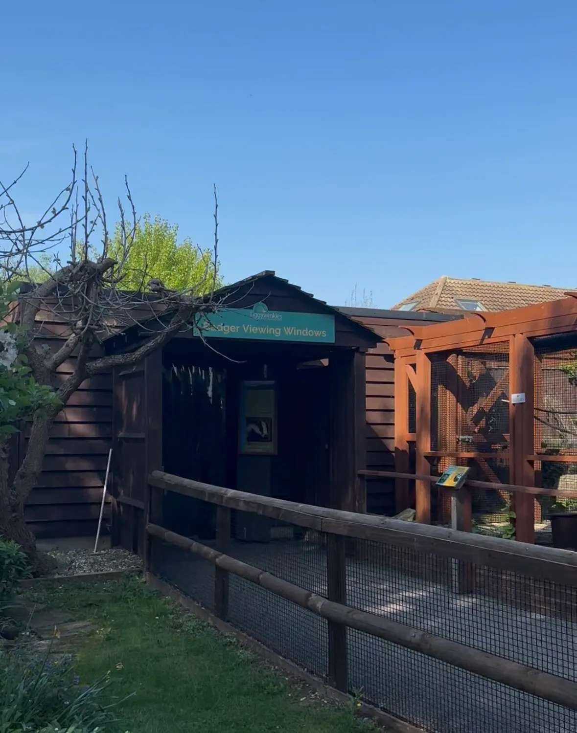 Rustic wooden building with viewing windows and an orange enclosed structure beside it, set in a garden under clear blue sky.