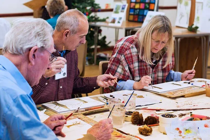 Three people of different ages engaged in a painting class, with brushes and watercolours at a table.