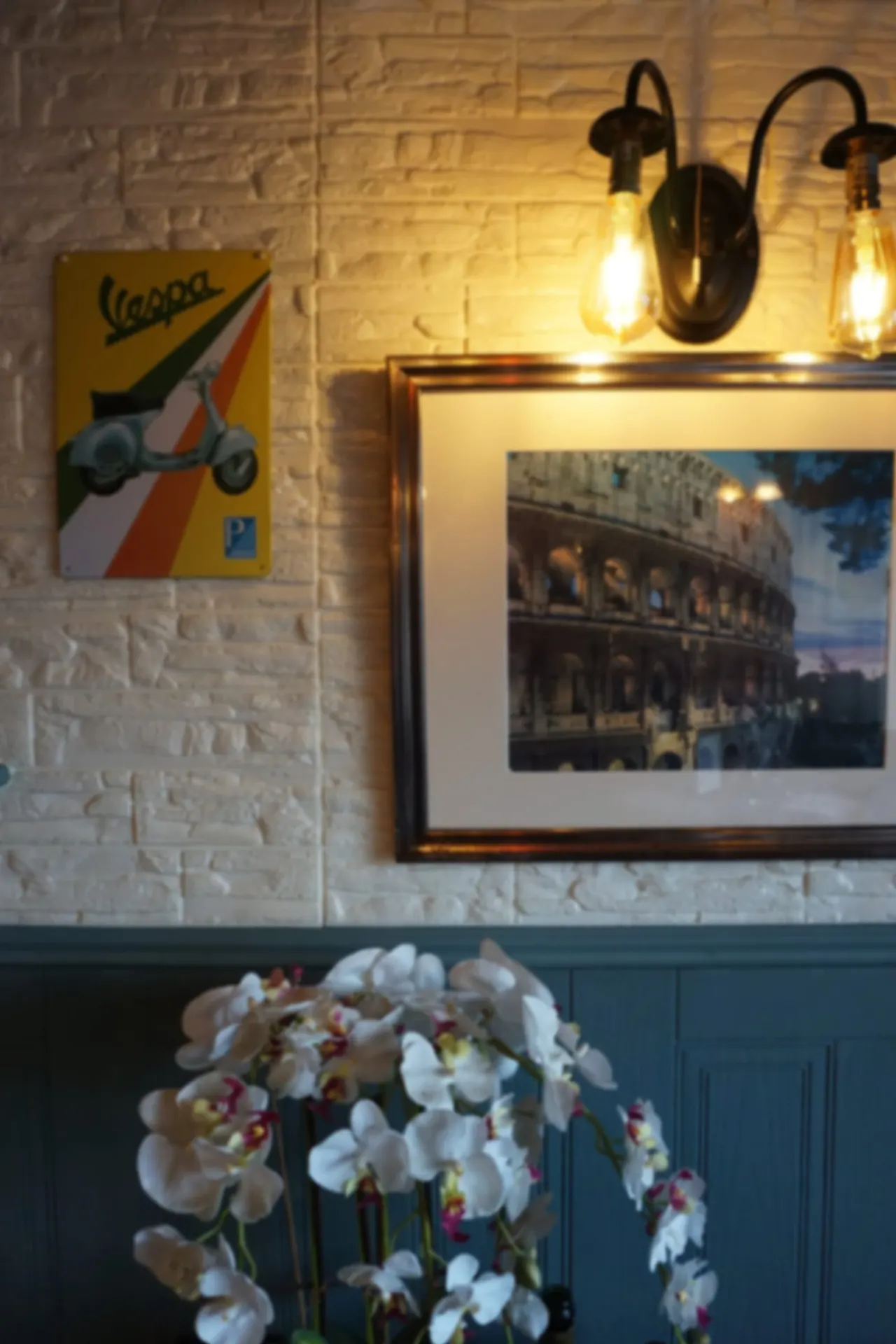White orchids in a vase sit beneath vintage Italian décor including a Vespa sign and framed architecture photograph on a brick wall.