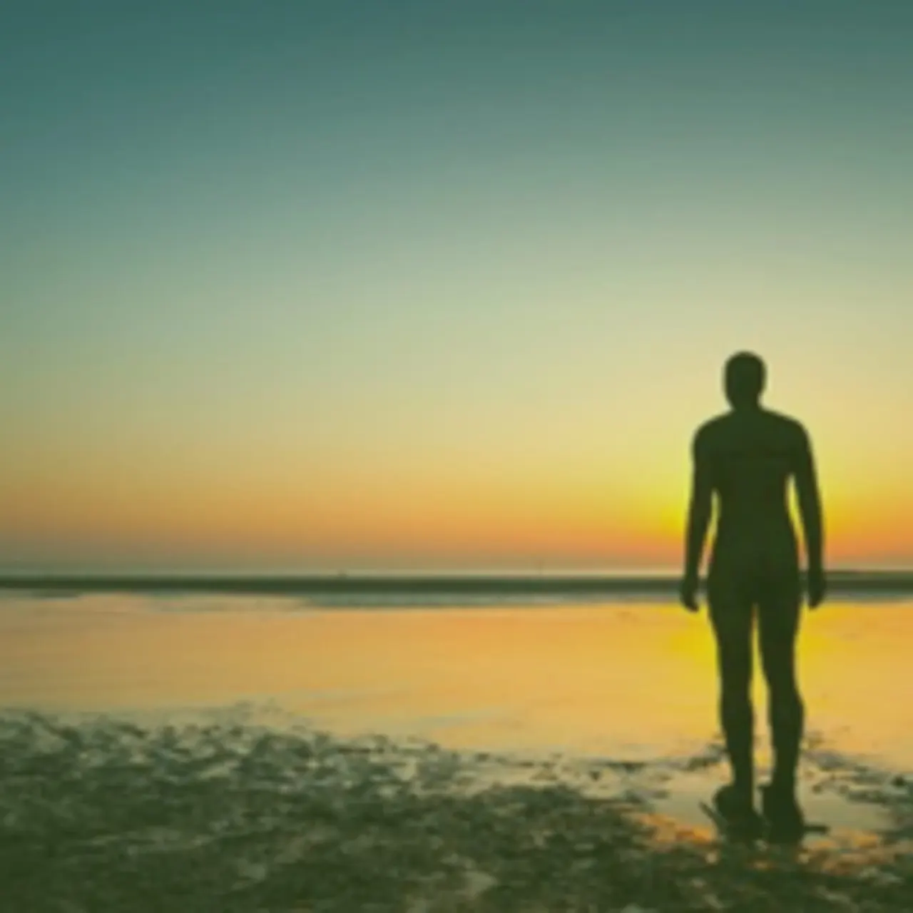 Person in wetsuit standing on beach at sunset looking towards the sea.