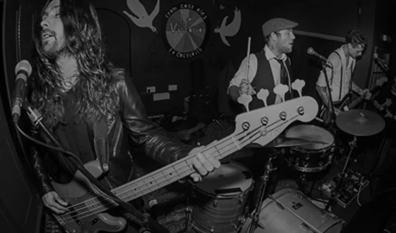 Black and white photograph of a four-piece rock band performing on stage at an indoor venue with decorative elements.