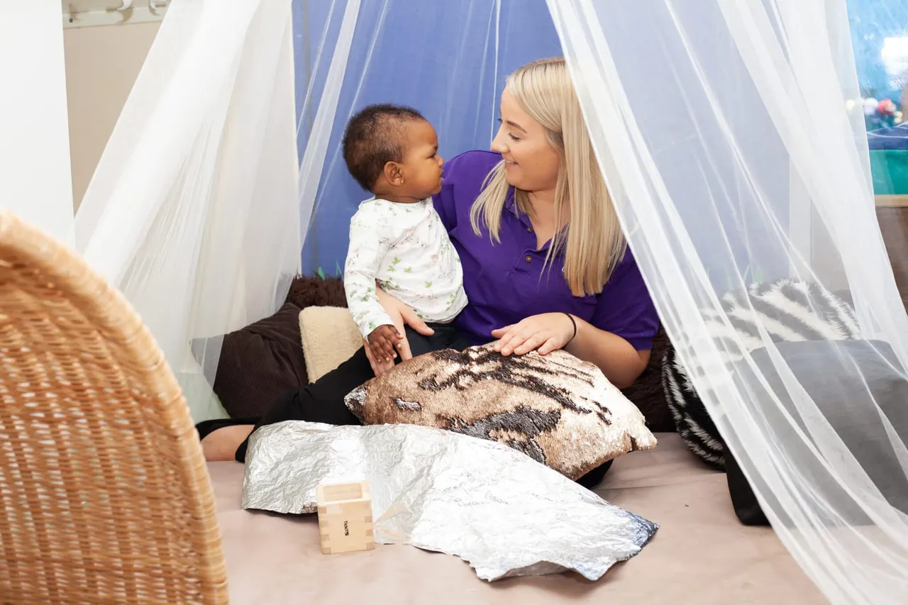 Adult carer and toddler playing together inside a sheer-draped sensory tent with cushions and textured fabrics.