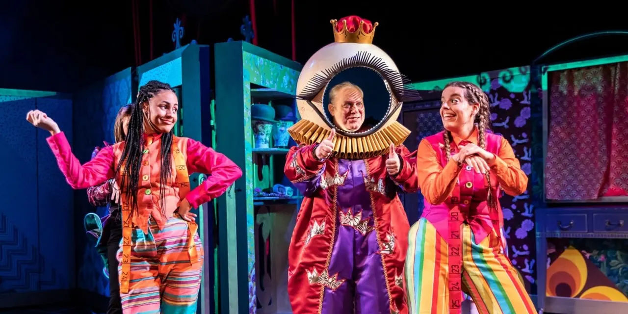 Three performers in colourful costumes on stage, with the centre performer wearing an elaborate crown and oversized eye prop during a theatrical production.