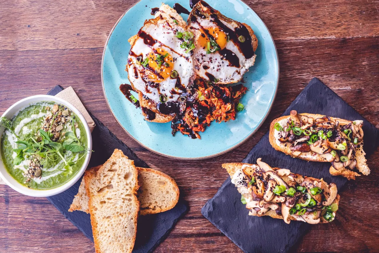 Overhead view of plated brunch dishes including creamy green soup, toasted bread slices, and multiple open-faced toasts topped with eggs, mushrooms and garnishes.