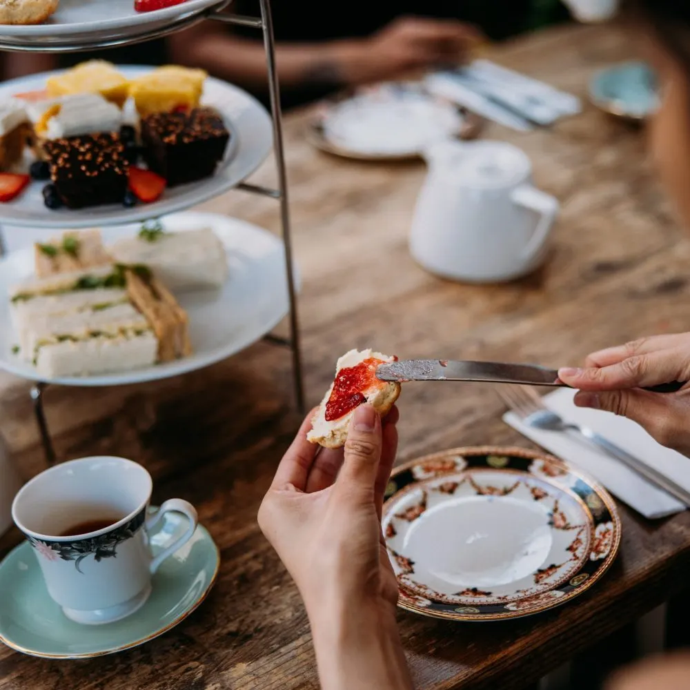 Person spreading jam on scone at tiered afternoon tea stand with cakes, sandwiches and tea.