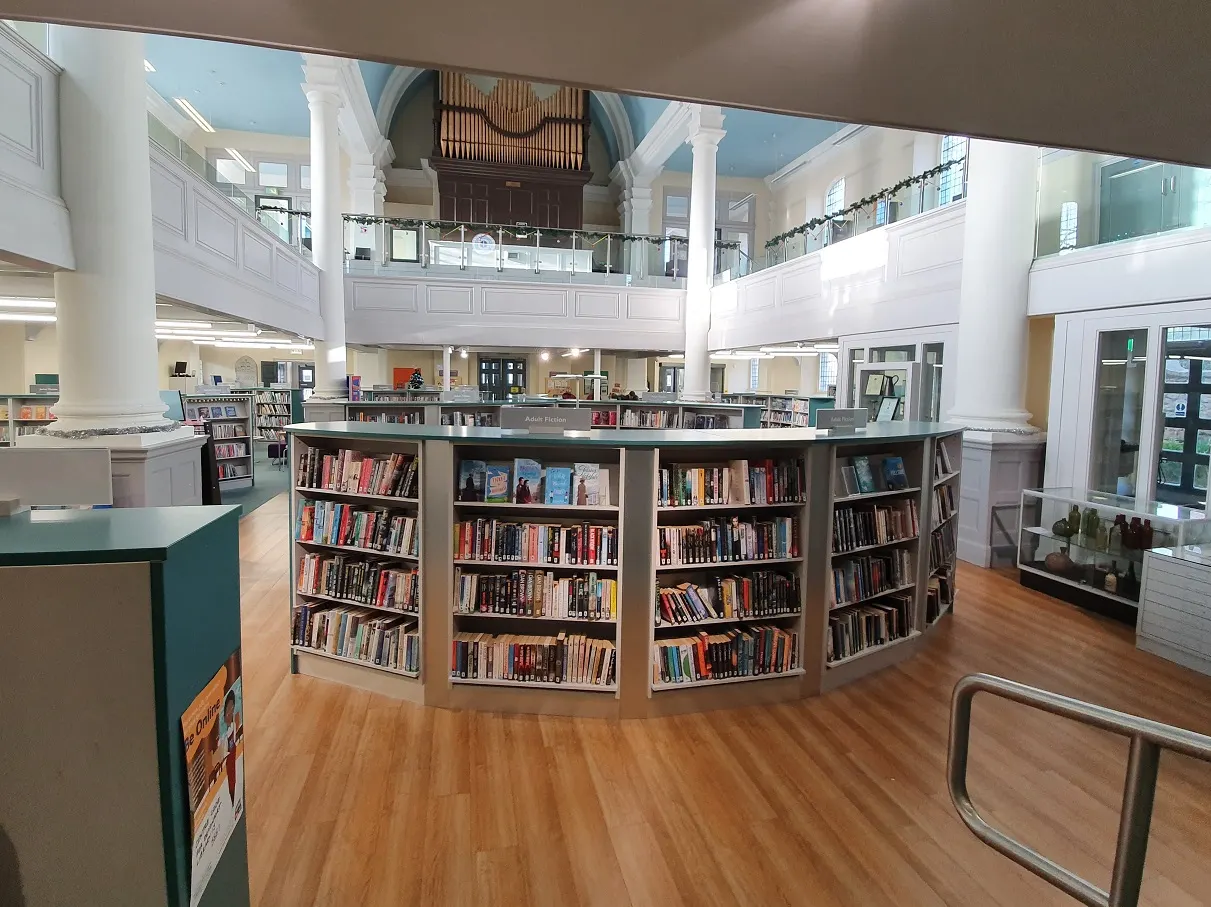 Interior of a bright, modern library with curved book shelves in the foreground, an organ visible on the upper gallery, and white columns throughout the spacious hall.
