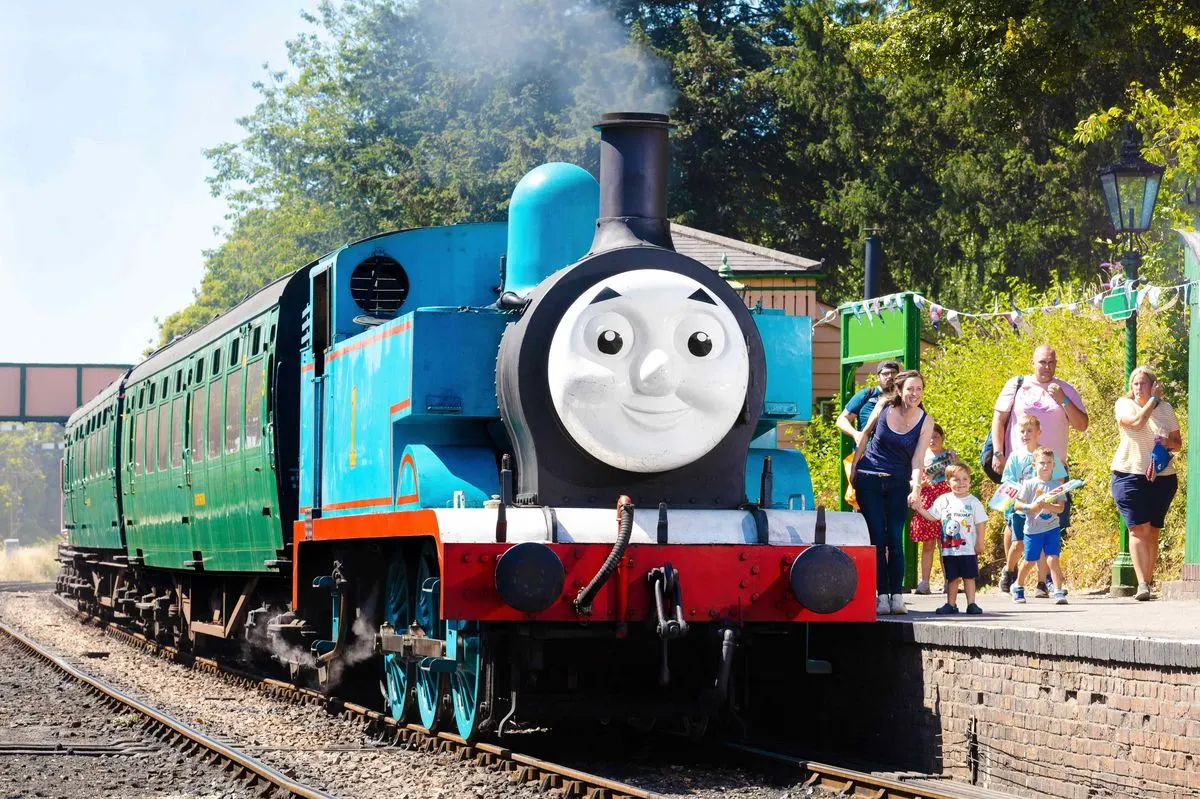 Cartoon-faced blue and red steam locomotive with passengers and families waiting on the platform beside the track.