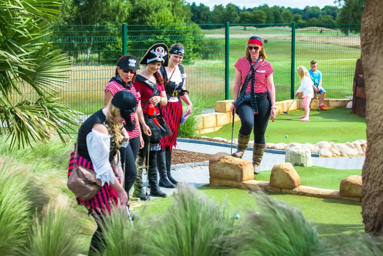 Adults and children in pirate fancy dress playing mini golf outdoors on a sunny day.
