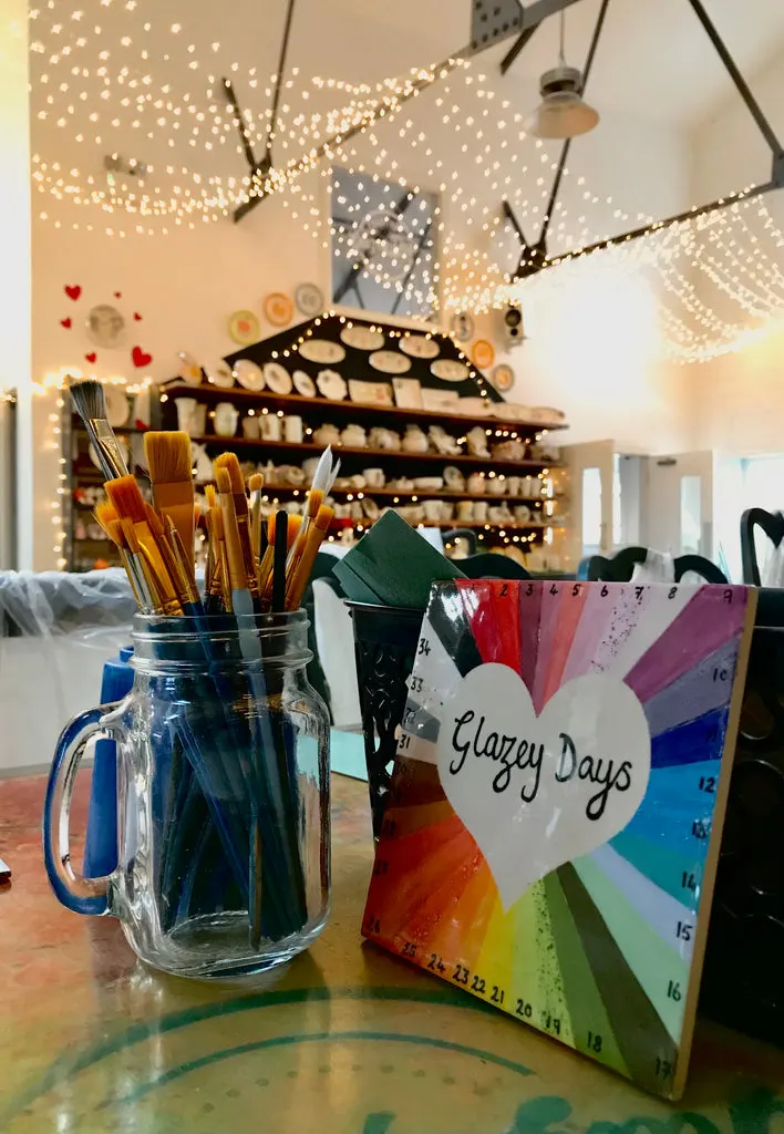 Art supplies and a colour wheel on a table inside a ceramics studio with shelves of pottery and hanging string lights.