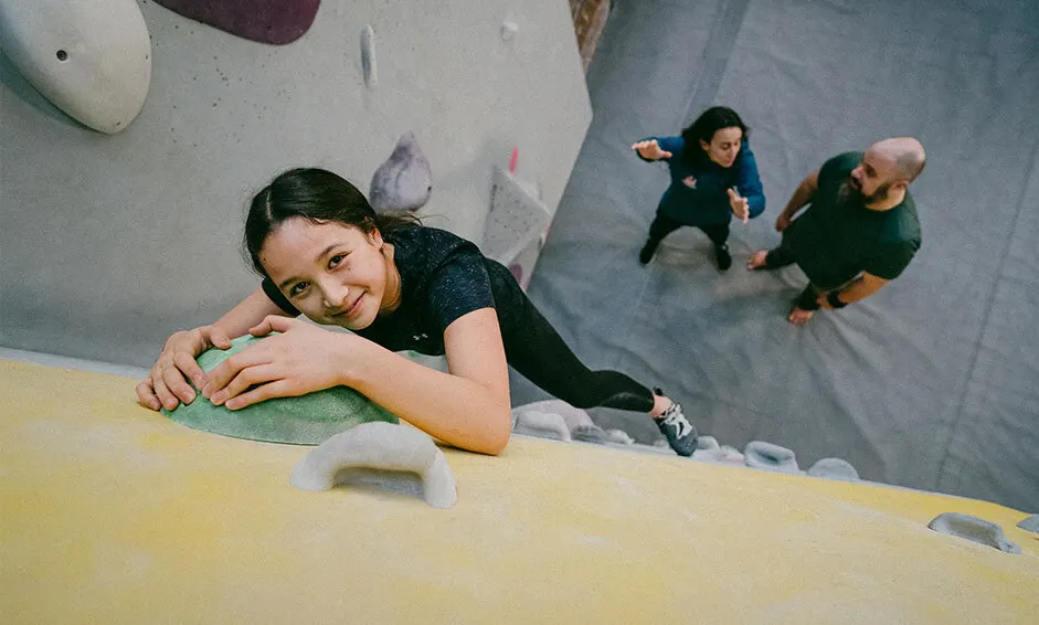 Woman climbing an indoor wall with handholds whilst two people watch from below.