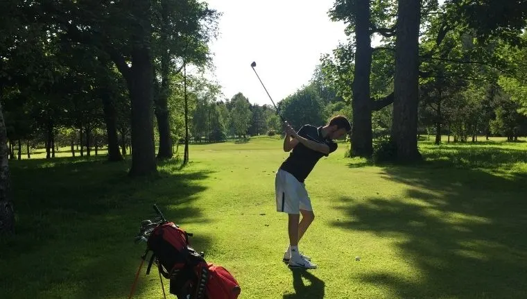 Golfer mid-swing on a tree-lined fairway with red golf bag in foreground.