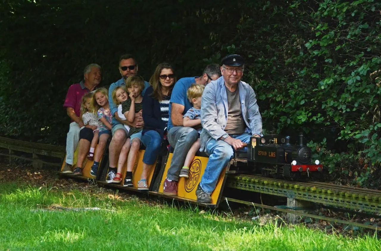 Multi-generational family group riding in a yellow open-sided carriage pulled by a miniature steam locomotive through tree-lined tracks.