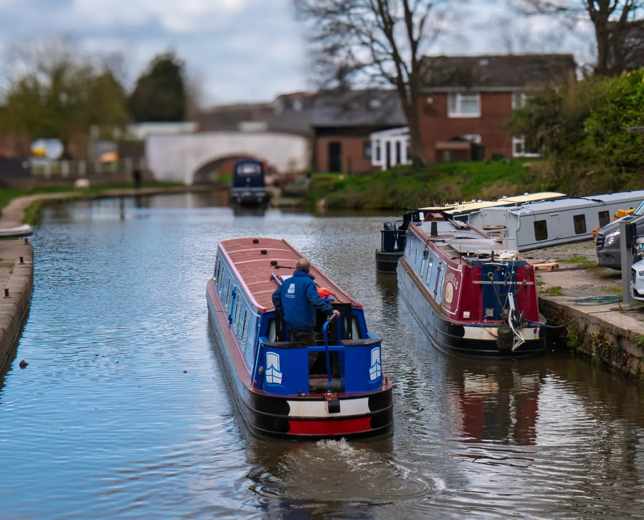 Narrowboat with two people aboard navigating a canal, with moored boats and brick buildings alongside the waterway.