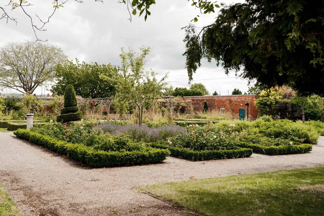 Formal garden with gravelled pathways, hedged borders, lavender plants and wooden pergolas, with a brick building in the background.