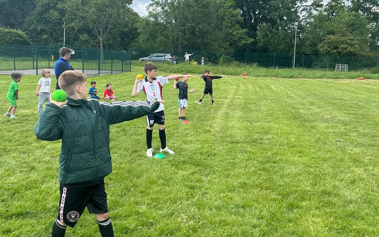 Children playing football and practising ball skills on an open grass pitch with an instructor supervising.