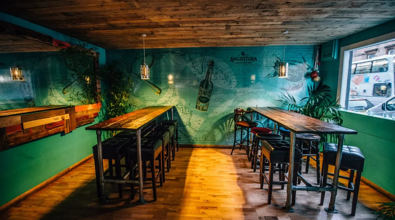 Empty bar interior with high tables, bar stools, wooden ceiling, turquoise walls decorated with maps and hanging bottles.