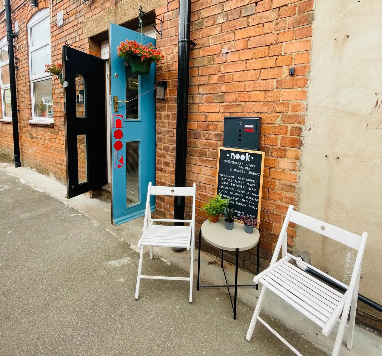 Exterior seating area with white folding chairs outside a brick building with a blue door and hanging red flowers.