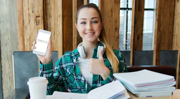 Young person at desk holding up a smartphone while thumbing towards it, with books and a cup nearby.