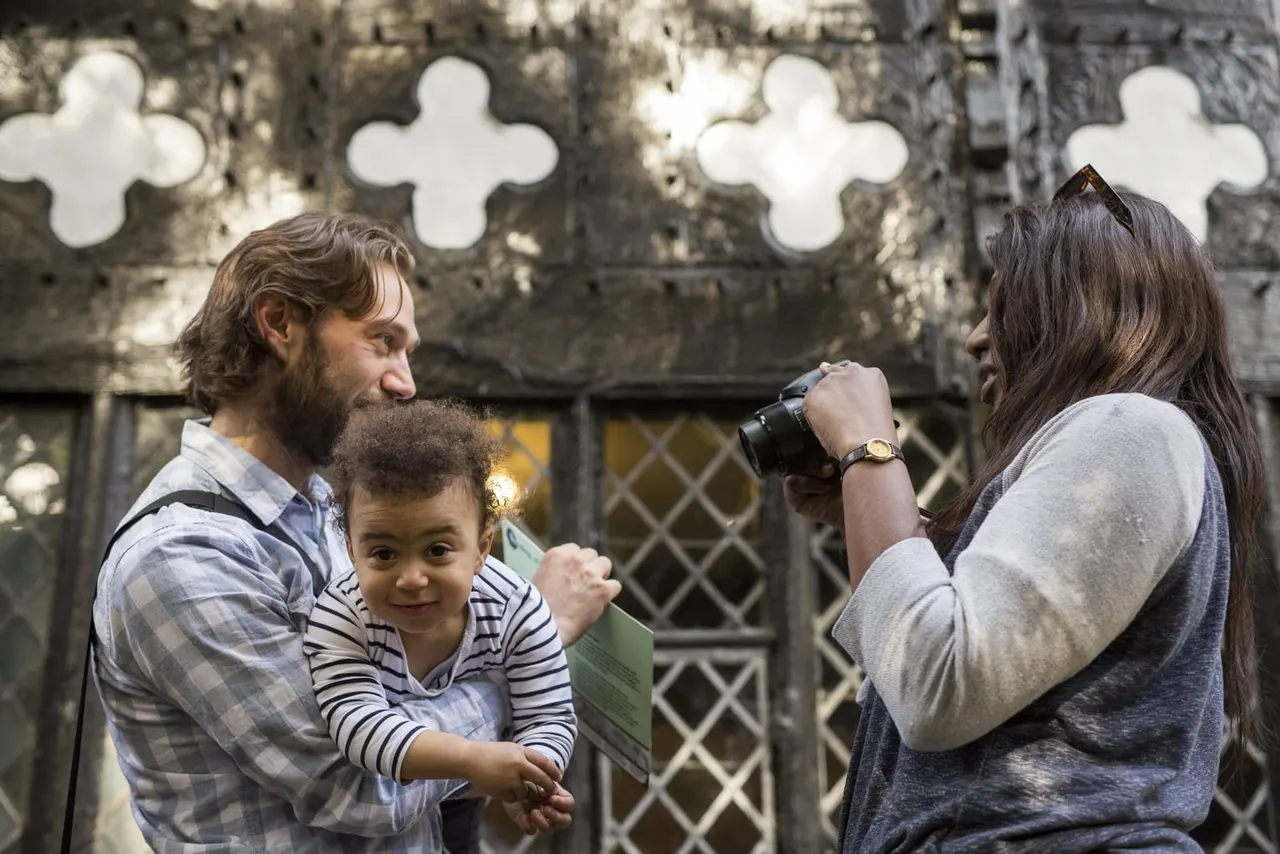 Man holding toddler posing whilst woman photographs them against ornate stone wall with quatrefoil patterns.