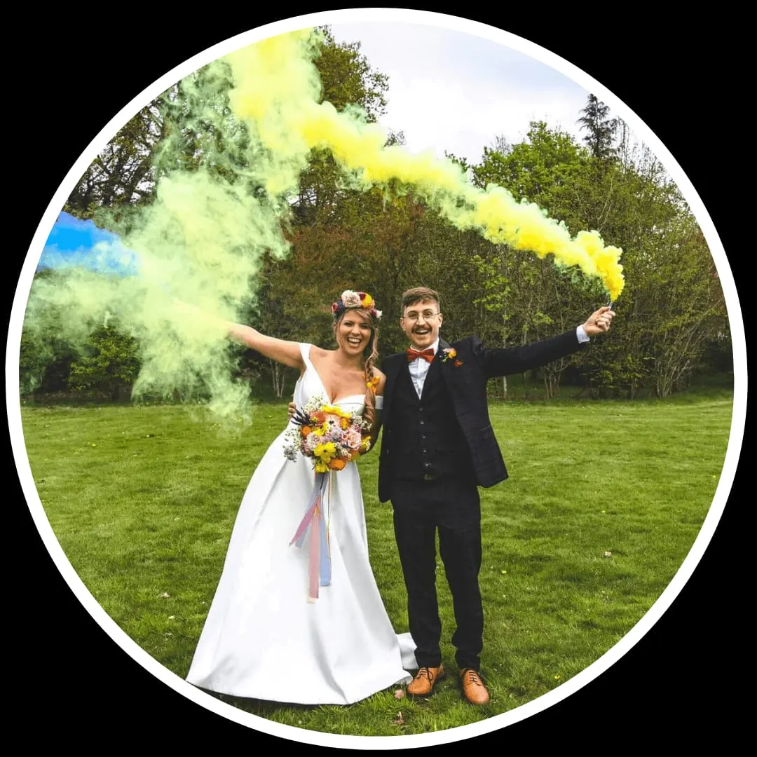 Bride and groom holding coloured smoke bombs in a grassy field surrounded by trees.
