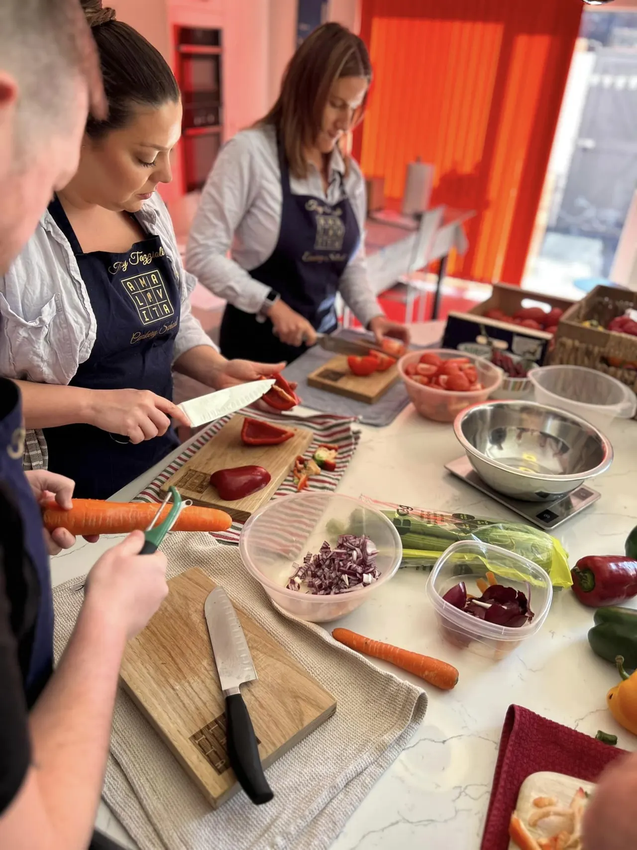 Several people in aprons prepare food together at a cooking class, chopping vegetables on wooden boards in a bright kitchen.
