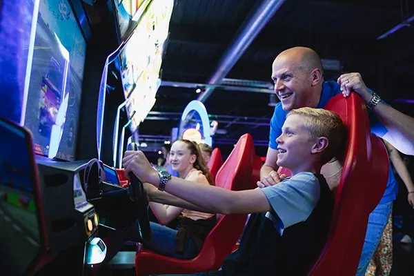 An adult and children play an arcade racing game together in a brightly lit gaming venue with red seats.