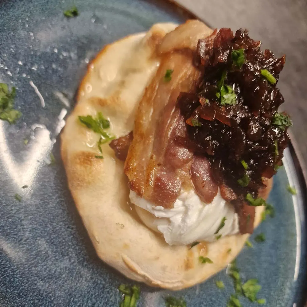 A toasted baguette filled with crispy bacon, white cheese and dark leafy greens, garnished with chopped parsley.