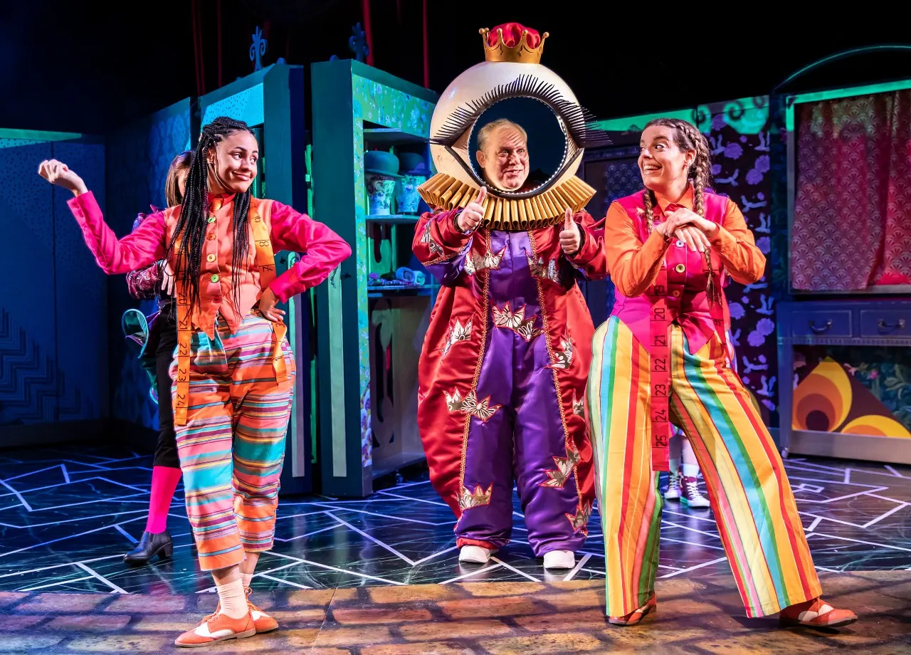 Three performers in colourful costumes perform on a theatre stage with vibrant set design and lighting.