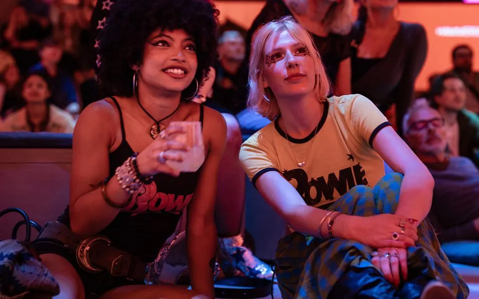 Two women sitting together enjoying entertainment in a dimly lit venue with coloured lighting and other guests visible in the background.