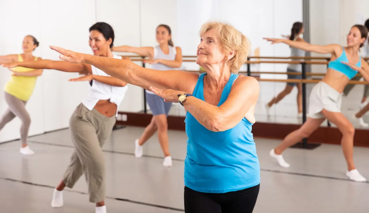 Group of women of mixed ages performing an aerobics or dance fitness class in a studio with a mirror wall.