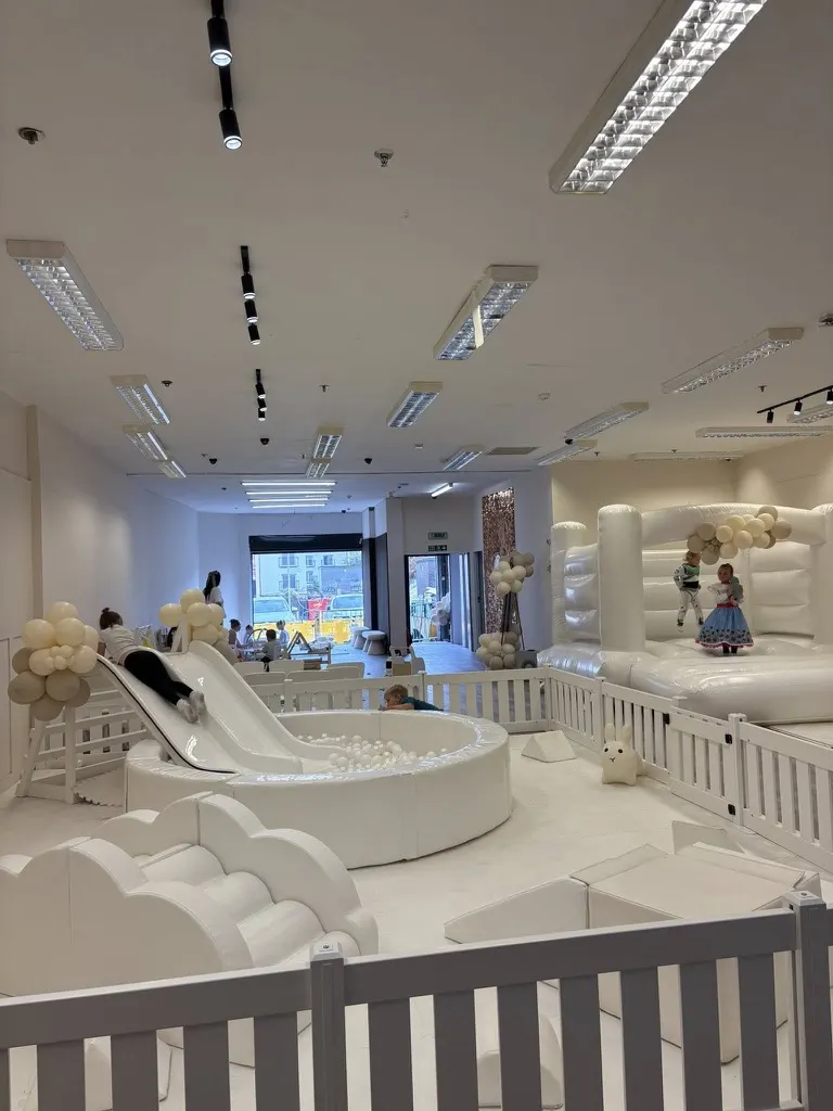 Modern indoor play centre with white soft play structures, slide, ball pit, and bouncy castle, children playing.