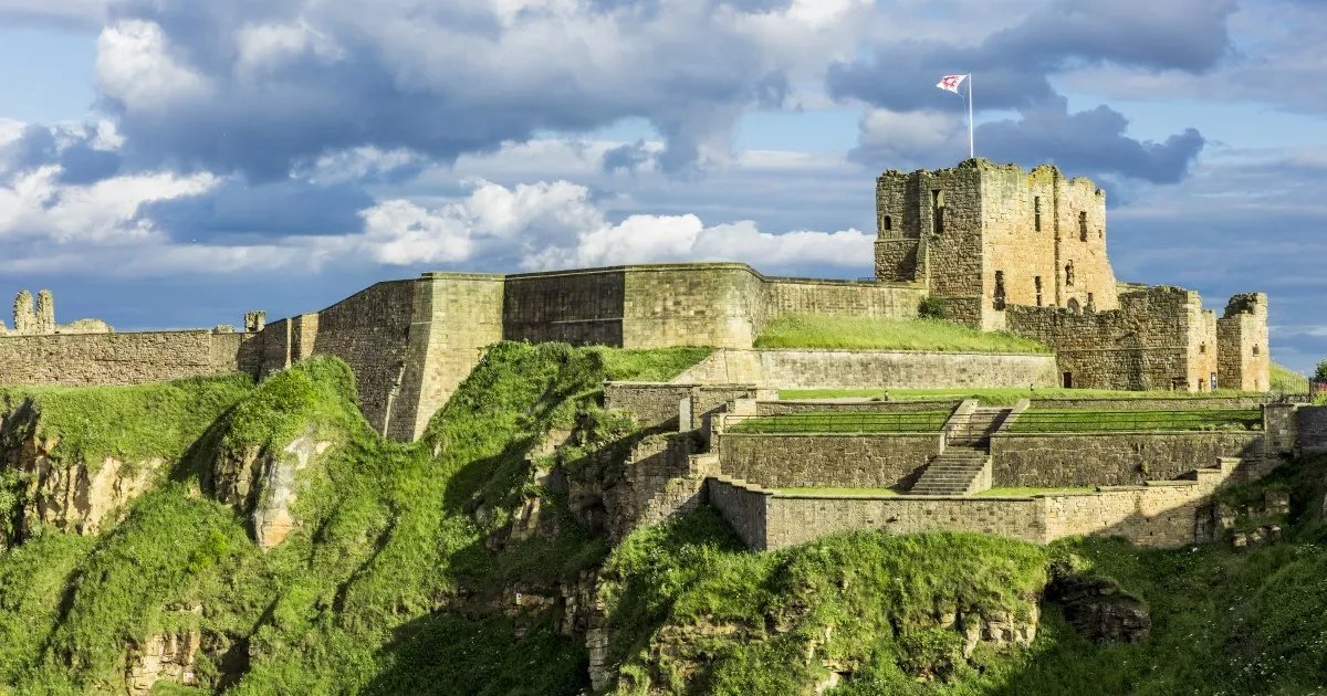 Medieval stone castle ruins on a cliff top with flag flying, surrounded by ivy-covered walls under a cloudy sky.