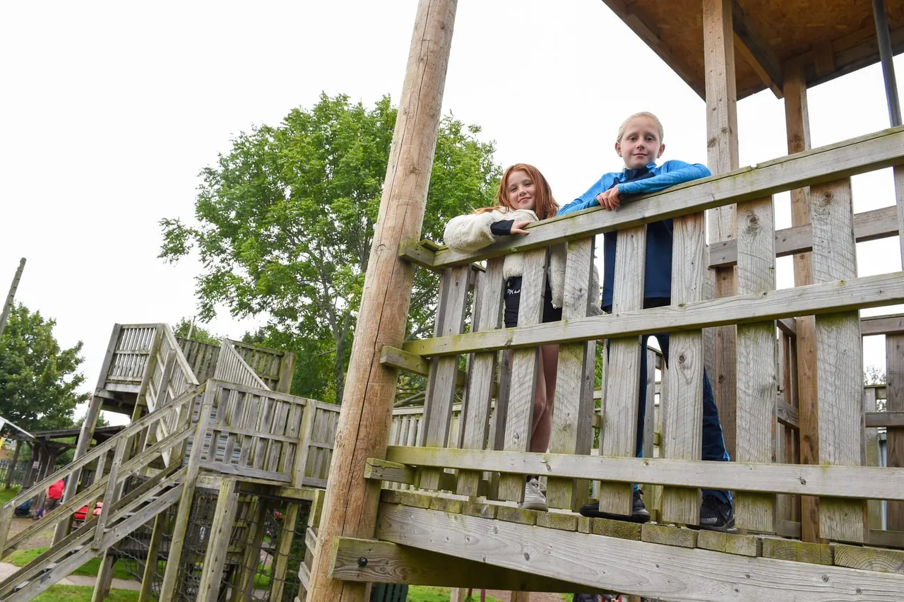 Two children standing on a wooden play structure with railings and lookout platforms in a treed outdoor play area.