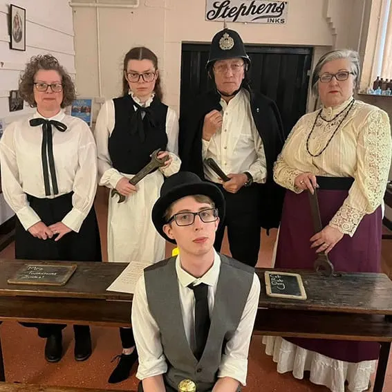 Five people in period costume stand in a vintage printing shop setting beneath a "Stephens Inks" sign.