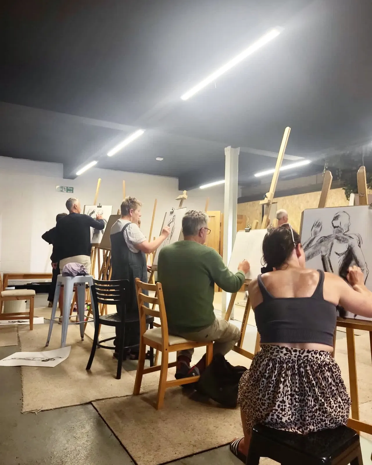 A group of adults drawing at easels during a life drawing class in a studio with bright overhead lighting.