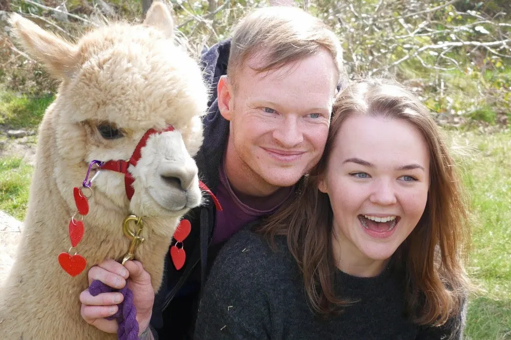 Two young people pose outdoors with a cream-coloured alpaca wearing a red halter and heart-shaped decorations.