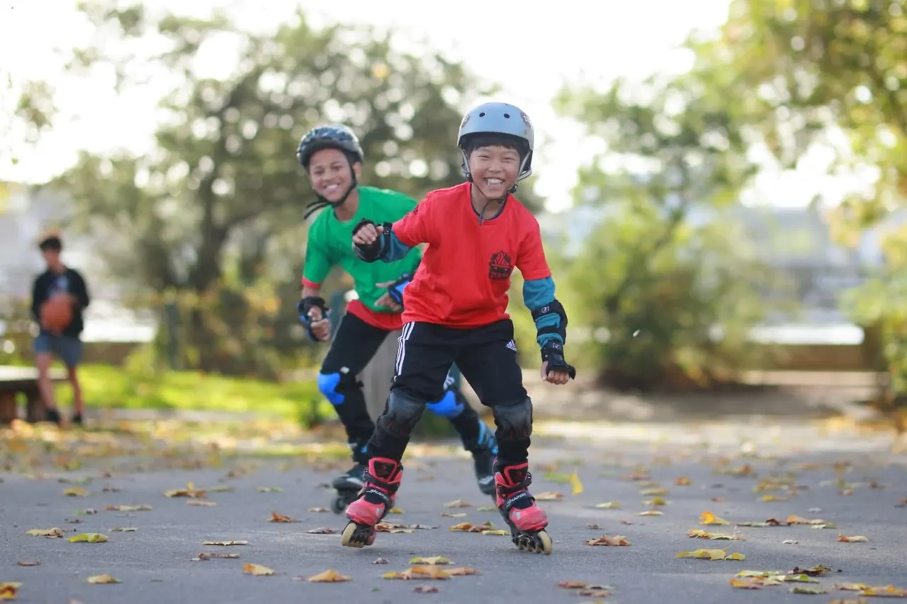 Two children wearing protective helmets and pads rollerblade together on a tree-lined path.