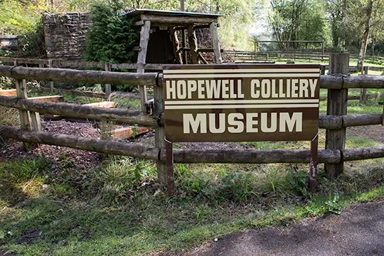 Wooden sign for Hopewell Colliery Museum with historic mining structures visible behind it in a wooded setting.