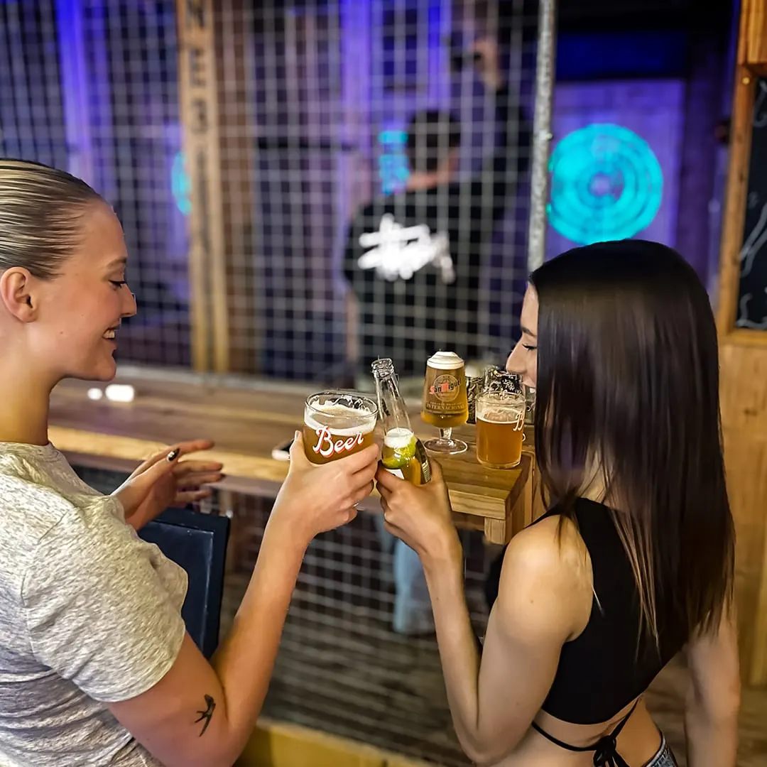 Two women toasting with beer bottles in front of an illuminated cityscape backdrop.