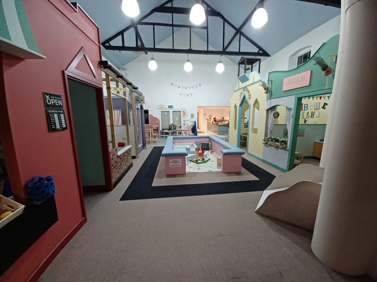 Indoor play space with colourful role-play buildings, a blue runway, soft seating, and overhead lighting for young children.