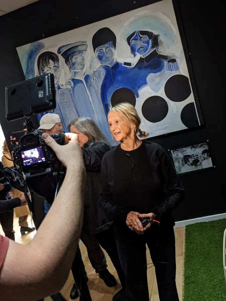 A woman being filmed by a cameraman in front of a blue and white mural depicting figures in military uniforms.