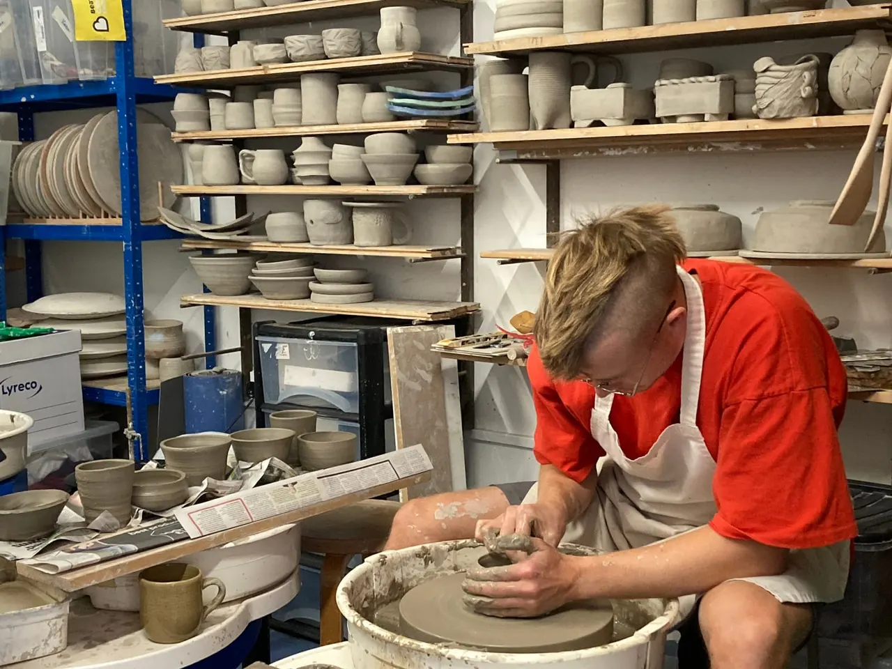 A person in a red jacket and apron hand-throws clay on a pottery wheel in a studio surrounded by shelves of finished ceramic pieces.