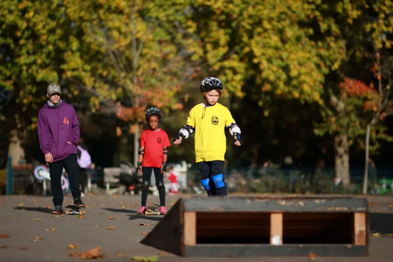 Children skateboarding and rollerblading at a park with autumn foliage in the background.