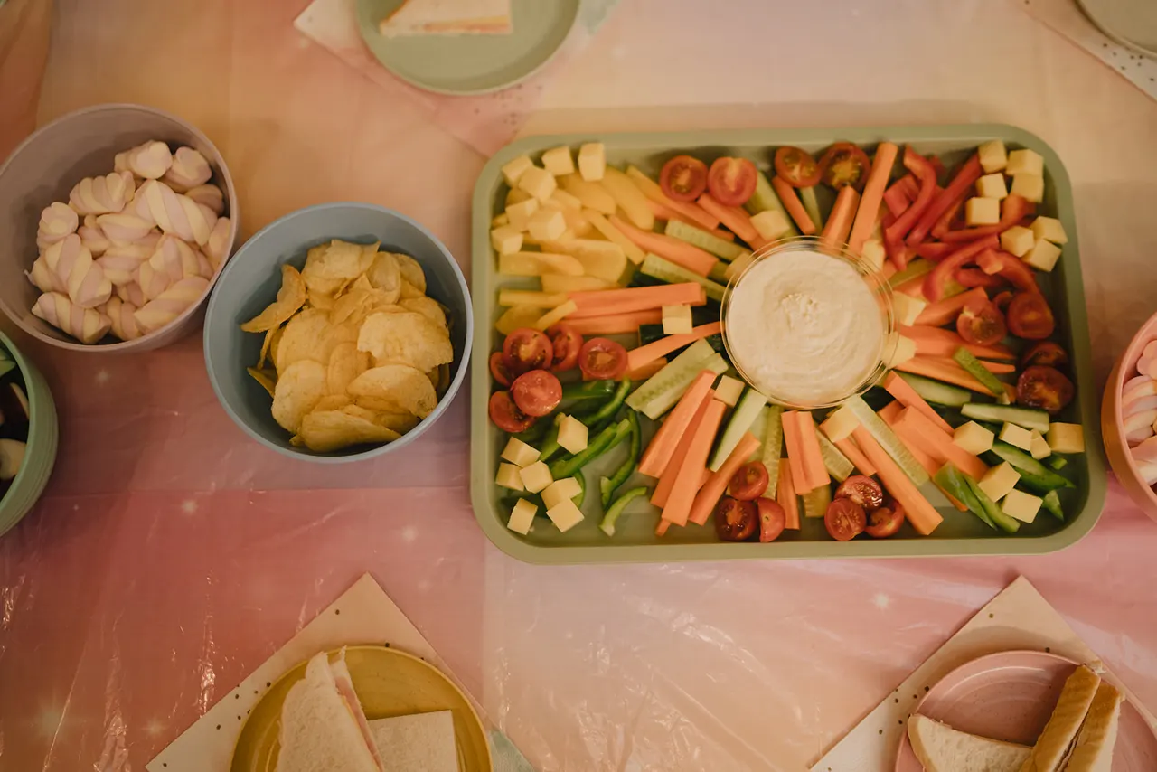 A spread of party food including a vegetable platter with dip, crisps, sliced onions, and cheese on a pink surface.