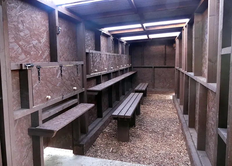 Interior of a farm animal shelter with wooden benches, feeding rails and gravel flooring.